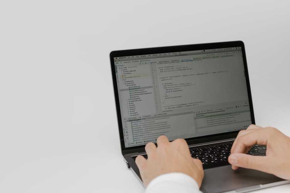 A person typing on a laptop with a black keyboard, seated at a desk against a plain white background. The laptop screen displays a code editor with multiple lines of programming code and a project dir
