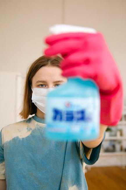 A person dressed in full protective hazmat gear, including a white suit, gloves, face mask, and goggles, standing in a modern kitchen with light-colored wooden wall panels and dark upper cabinets. The